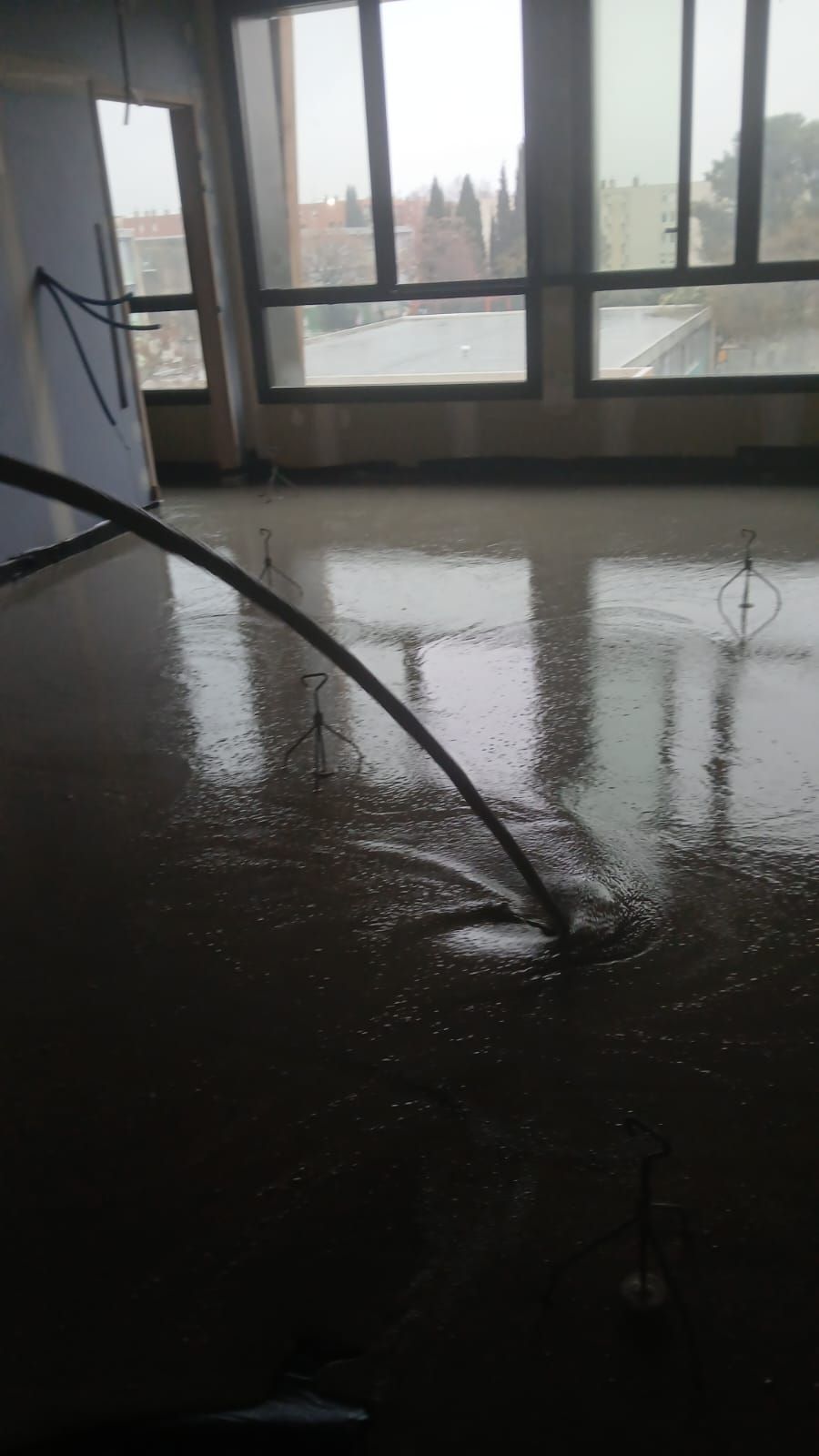 Un tuyau déverse du béton liquide sur le sol d'une pièce en construction dotée de grandes fenêtres.