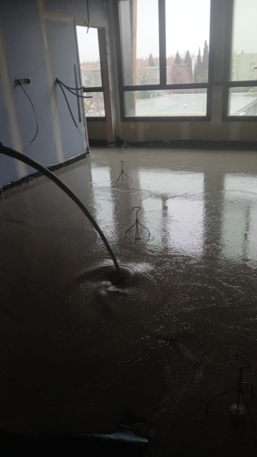 Un tuyau déverse du ciment liquide sur le sol d'un chantier de construction intérieur doté de grandes fenêtres.