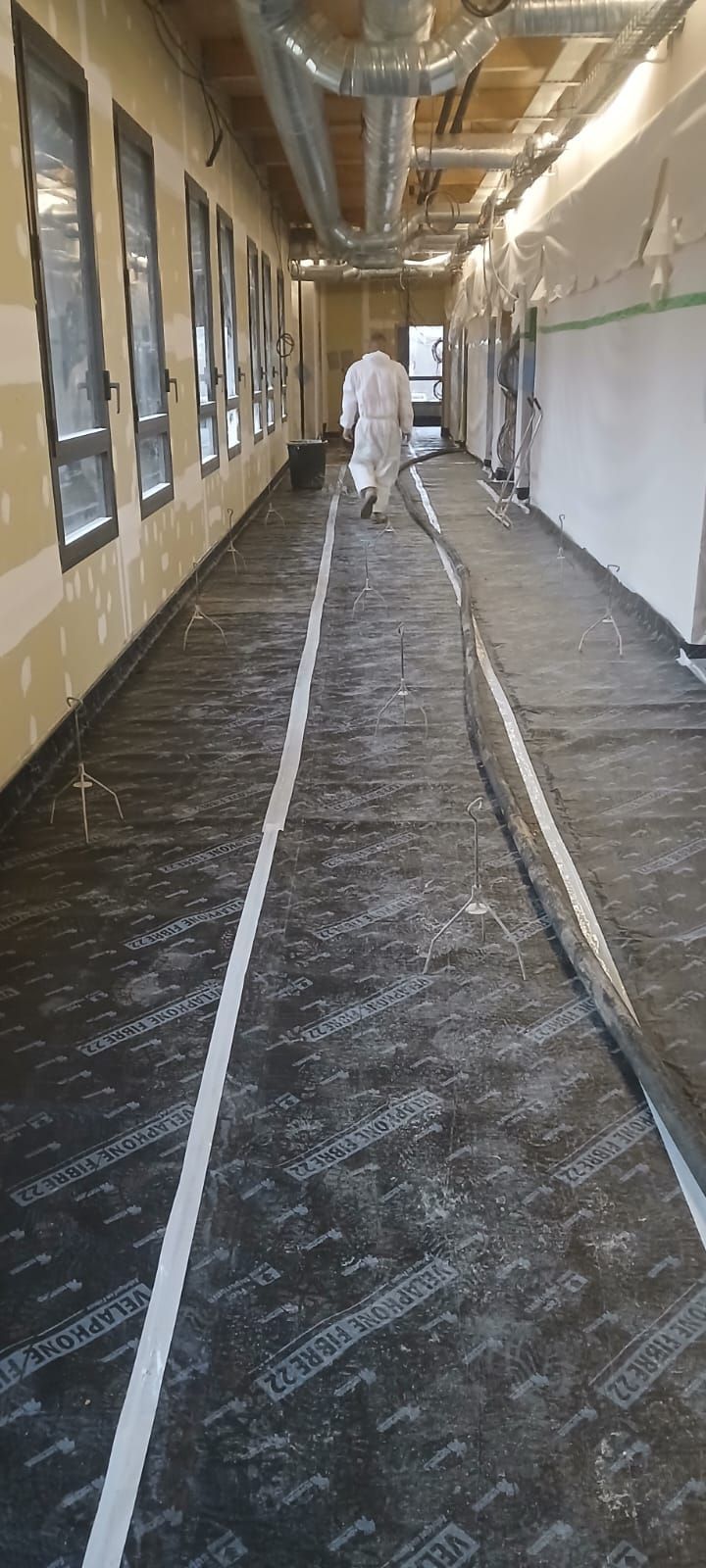 Une personne en tenue de protection marche dans un couloir de chantier dont le sol est recouvert d'une sous-couche noire et les conduits de plafond sont apparents.