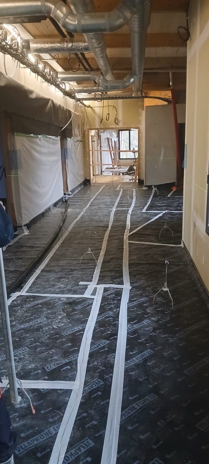 Couloir de chantier avec conduits métalliques apparents au plafond et ruban adhésif blanc marquant le plan au sol sur une sous-couche noire.