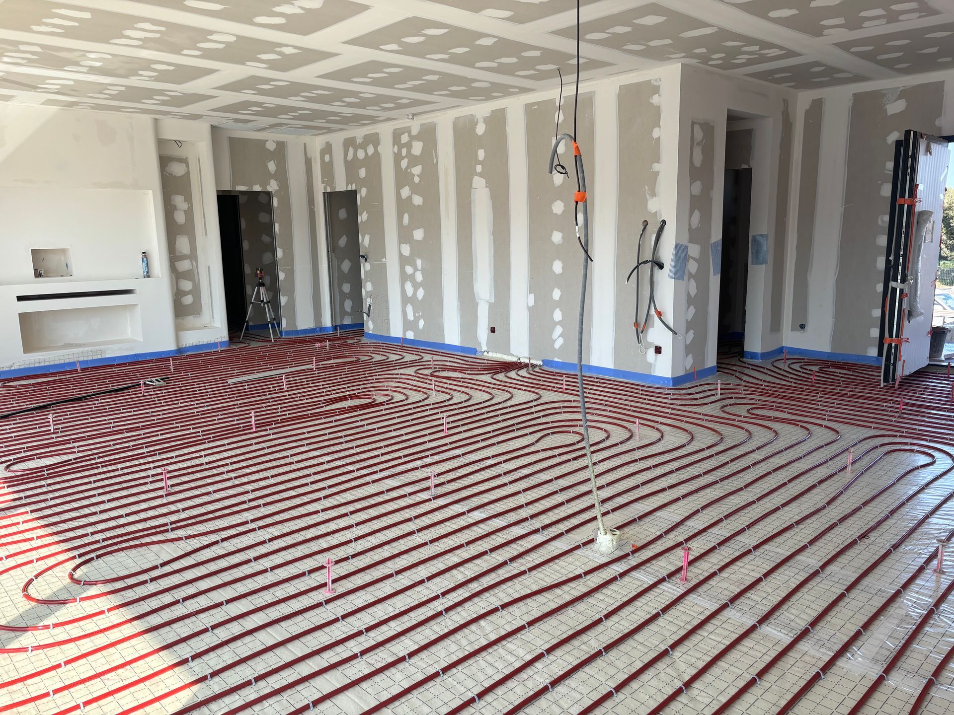 Chantier de construction intérieur avec des tuyaux de chauffage au sol radiant rouges installés sur un plancher sous les plaques de plâtre du plafond et des murs.