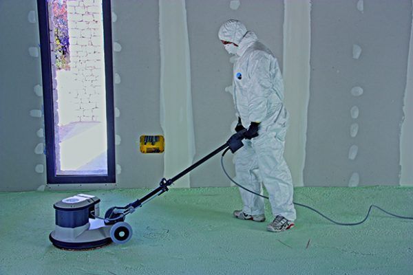 Une personne portant une combinaison de protection utilise une ponceuse à parquet sur une surface verte à l'intérieur d'une pièce aux murs en plaques de plâtre non finies.