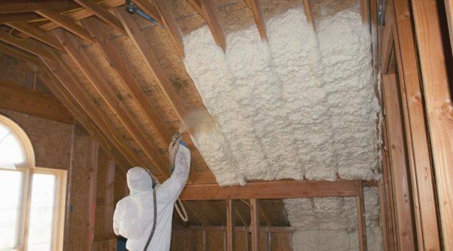 Un ouvrier en tenue de protection utilise un embout de pulvérisation pour appliquer de la mousse isolante blanche entre les chevrons en bois d'un grenier.