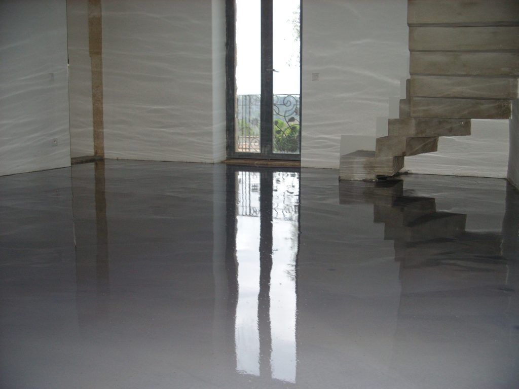 Une pièce moderne avec un sol gris lisse et brillant reflétant une haute fenêtre et un escalier en béton adjacent.