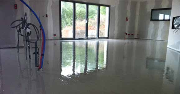 Une surface de sol lisse, humide et fraîchement coulée, autonivelante, dans une pièce non finie avec de grandes portes vitrées et des câbles apparents.