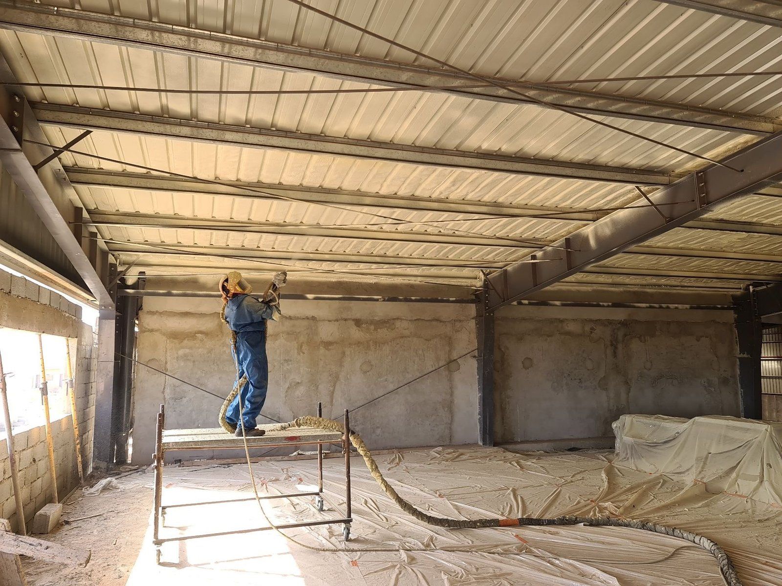 Un ouvrier en combinaison bleue pulvérise de l'isolant en mousse sur le plafond en tôle ondulée de l'intérieur d'un bâtiment en construction.