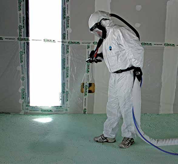 Une personne vêtue d'une tenue de protection blanche intégrale et portant un masque respiratoire pulvérise de l'isolant en mousse sur un sol dans une zone de construction délimitée par du ruban adhésif.
