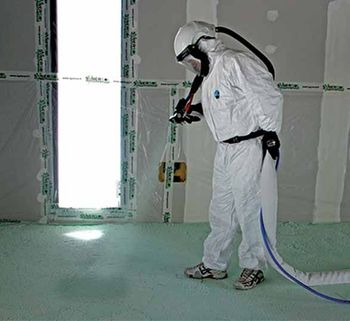 Un ouvrier vêtu d'une combinaison de protection blanche et d'un casque de protection pulvérise de l'isolant sur le sol d'une pièce à l'intérieur d'une zone de travail scellée.