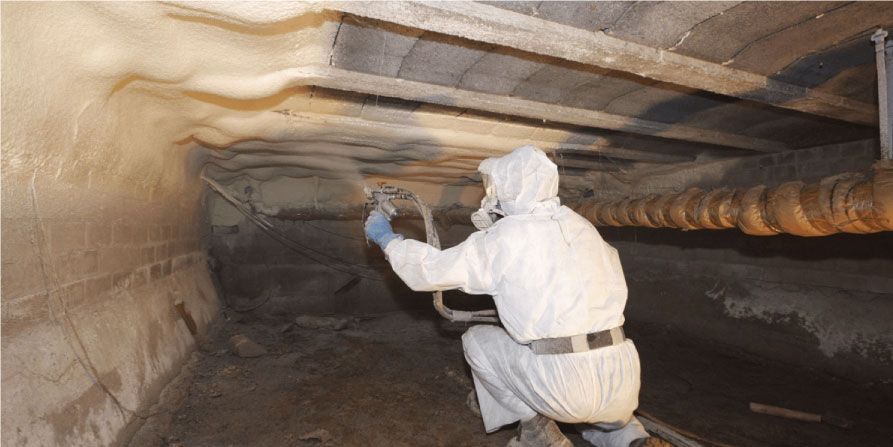 Un ouvrier portant un équipement de protection applique de l'isolant en mousse pulvérisée sur la face inférieure du plafond d'un vide sanitaire.