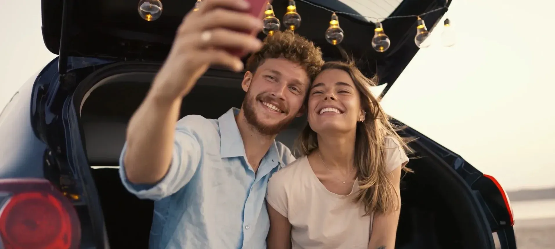 Una pareja sonríe y se toma una selfie junto al maletero abierto de un coche con globos al atardecer.