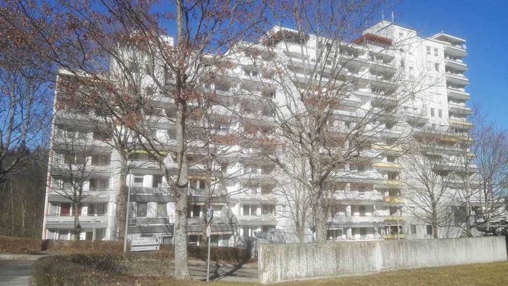 Mehrstöckiges weißes Wohnhaus mit Balkonen, davor Bäume.