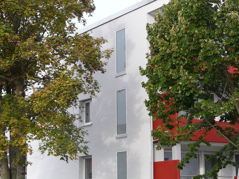 Weißes Gebäude mit hohen Fenstern und rotem Balkon, teilweise verdeckt von grünen und gelben Bäumen.