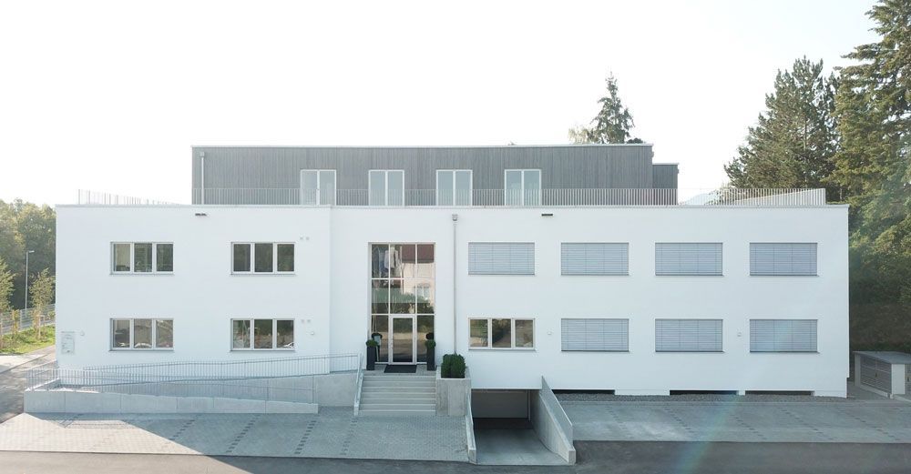 Weißes zweistöckiges Gebäude mit mehreren Fenstern, gläsernem Eingang und grauem Dach, im Freien gelegen.