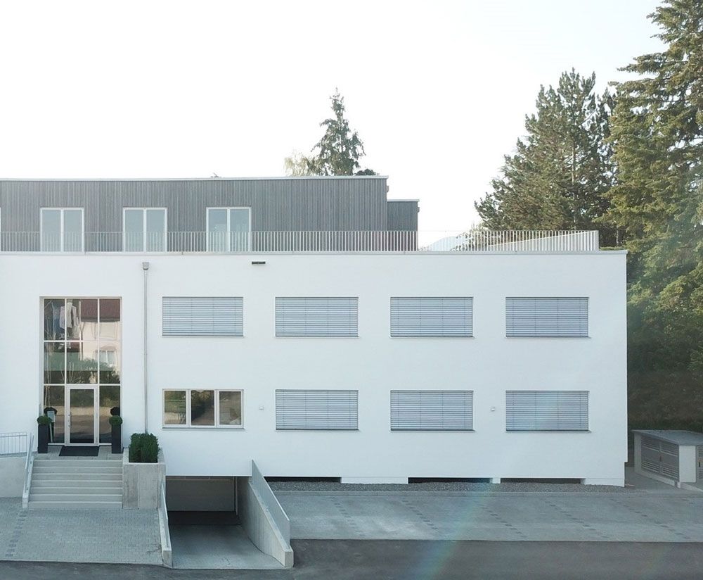 Weißes Gebäude mit mehreren Fenstern, Eingang mit Treppe und Garagentor.