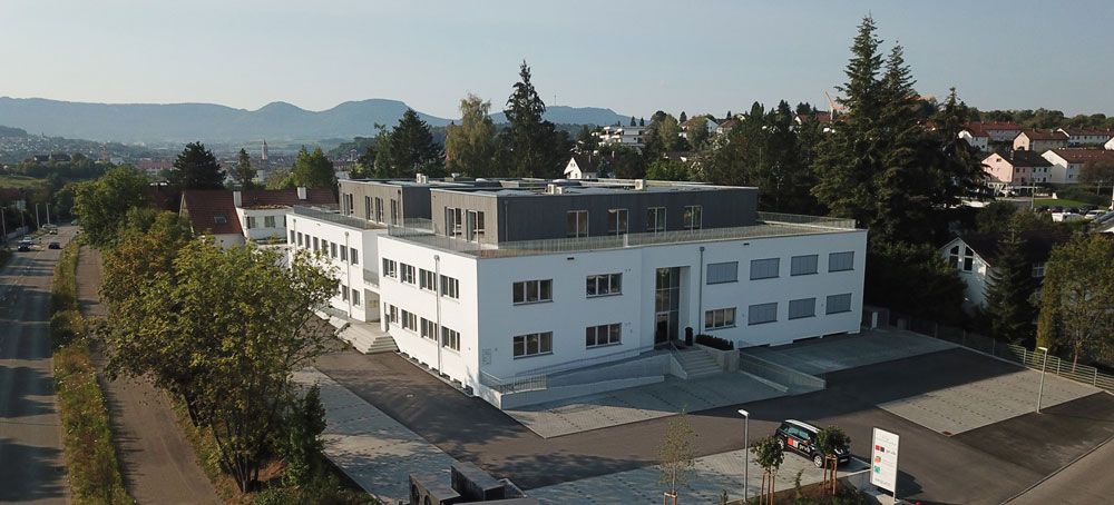 Luftaufnahme eines weißen Geschäftsgebäudes mit grauem Dach und Bäumen vor einer Bergkulisse.