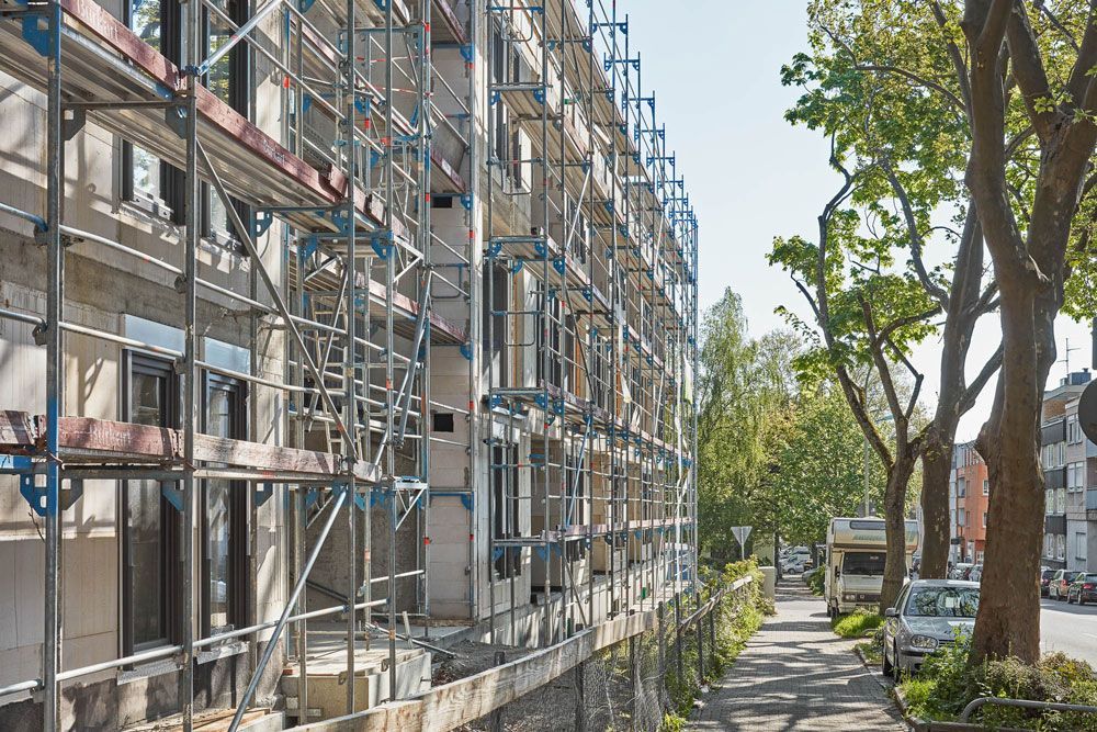 Die Gebäudefassade befindet sich im Bau und ist mit einem Gerüst versehen; Gehweg, Bäume und Straße sind sichtbar.