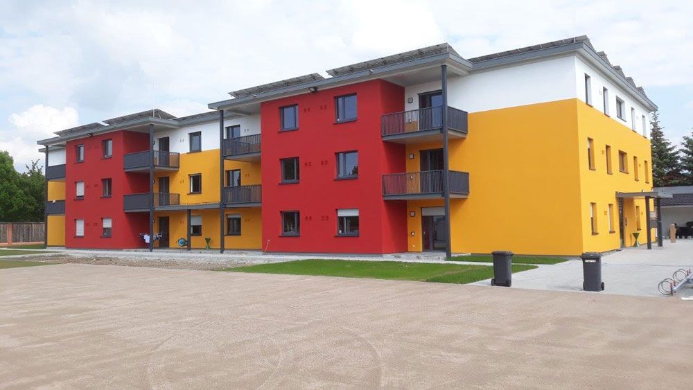 Buntes Wohnhaus mit roten, gelben und weißen Wänden, Balkonen und grauen Zierelementen unter einem bewölkten Himmel.