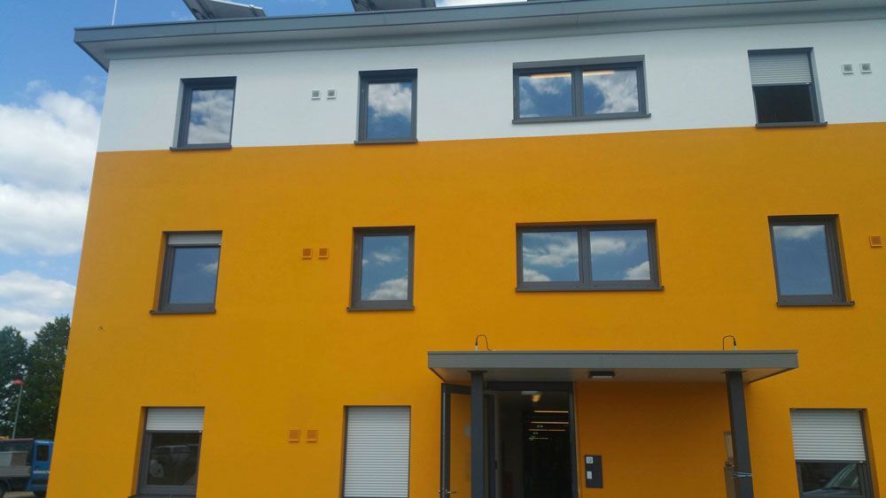 Gelb-weißes Gebäude mit dunkel gerahmten Fenstern, blauer Himmel im Hintergrund.