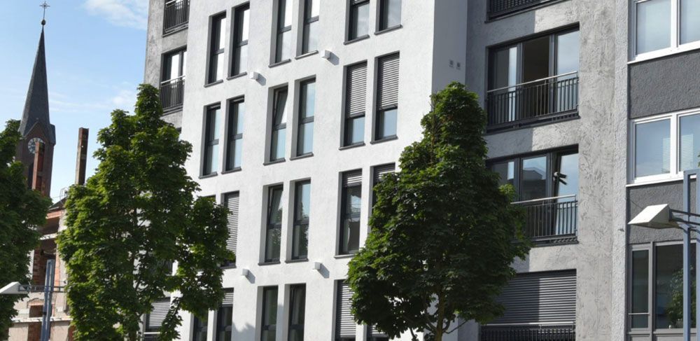 Modernes weißes Wohnhaus mit vielen Fenstern, Bäumen und einem Kirchturm im Hintergrund.