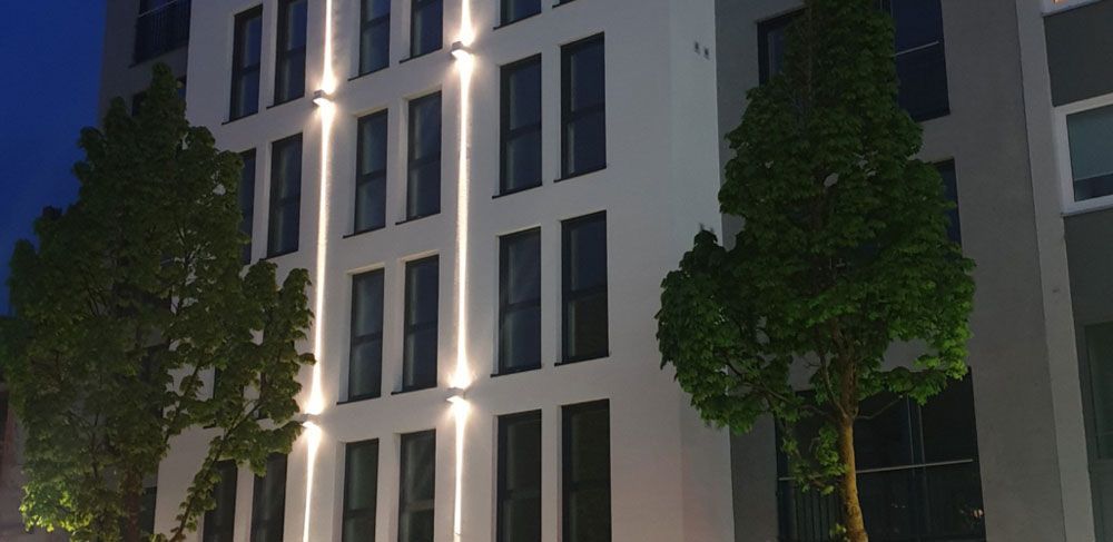 Modernes Gebäude mit vertikalen Außenleuchten, Fenstern und Bäumen, die in der Abenddämmerung beleuchtet werden.