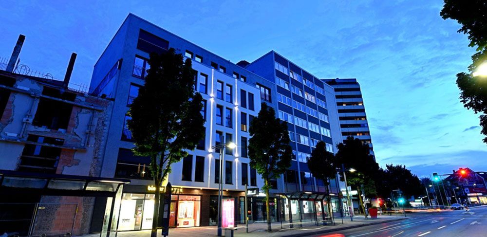 Ein mehrstöckiges Gebäude mit beleuchteten Fenstern an einer Stadtstraße in der Abenddämmerung.