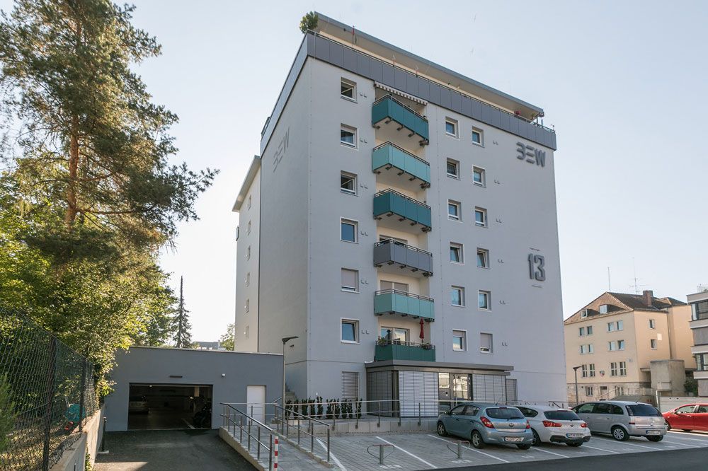 Mehrstöckiges Wohnhaus mit Balkonen, davor geparkten Autos und einer Garageneinfahrt.