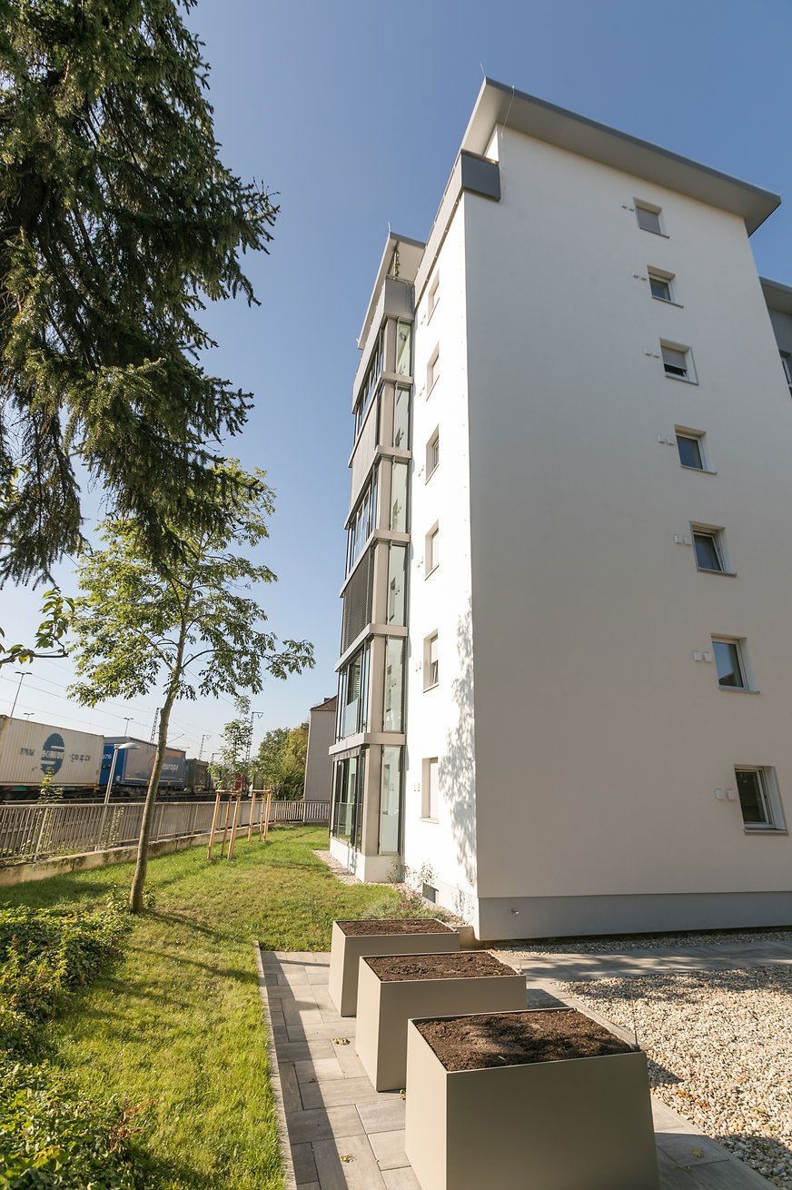 Modernes weißes Apartmentgebäude mit großen Fenstern, quadratischen Pflanzkästen und blauem Himmel.