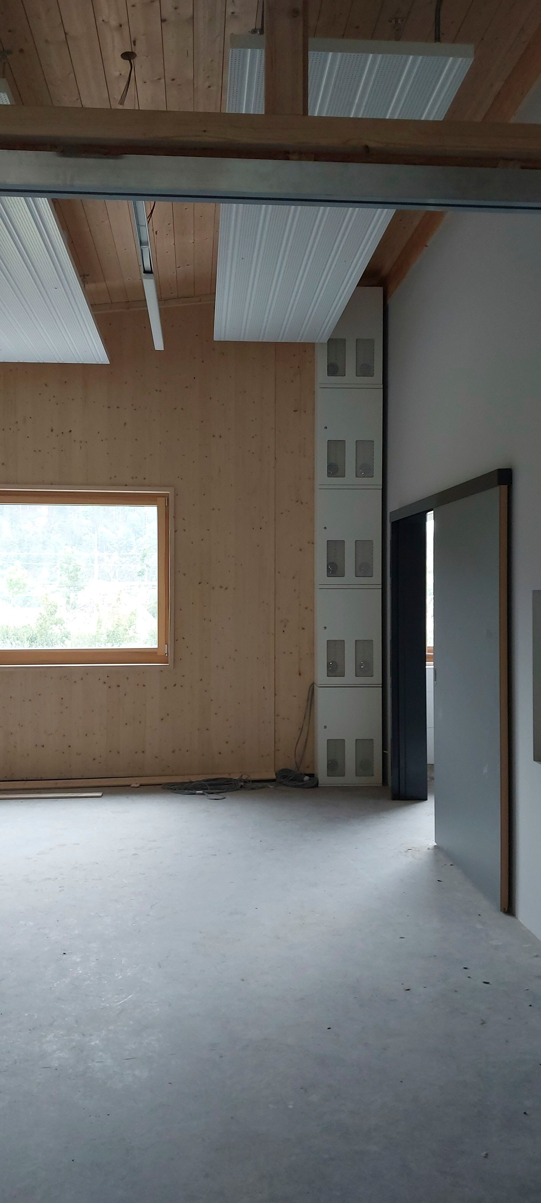 Innenansicht eines Zimmers mit unverputzten Holzwänden, einem Fenster und einer Tür. An einer Wand befinden sich weiße Aufbewahrungsschränke.