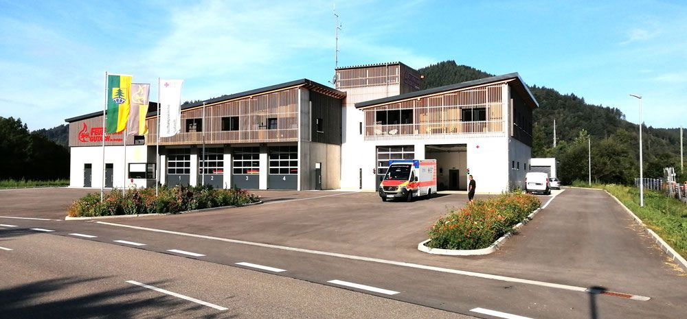 Feuerwache mit einem aus der Bucht fahrenden Krankenwagen. Grüne Flagge und Sträucher im Vordergrund.