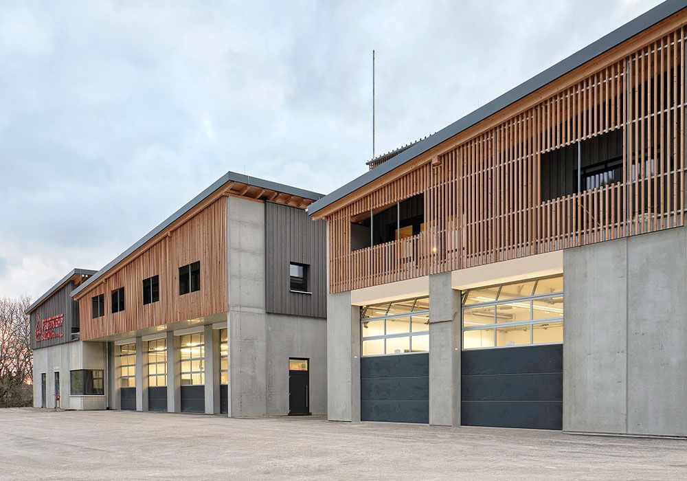 Moderne Feuerwache mit Fassade aus Beton und Holz, Garagentoren und Fenstern unter einem bewölkten Himmel.