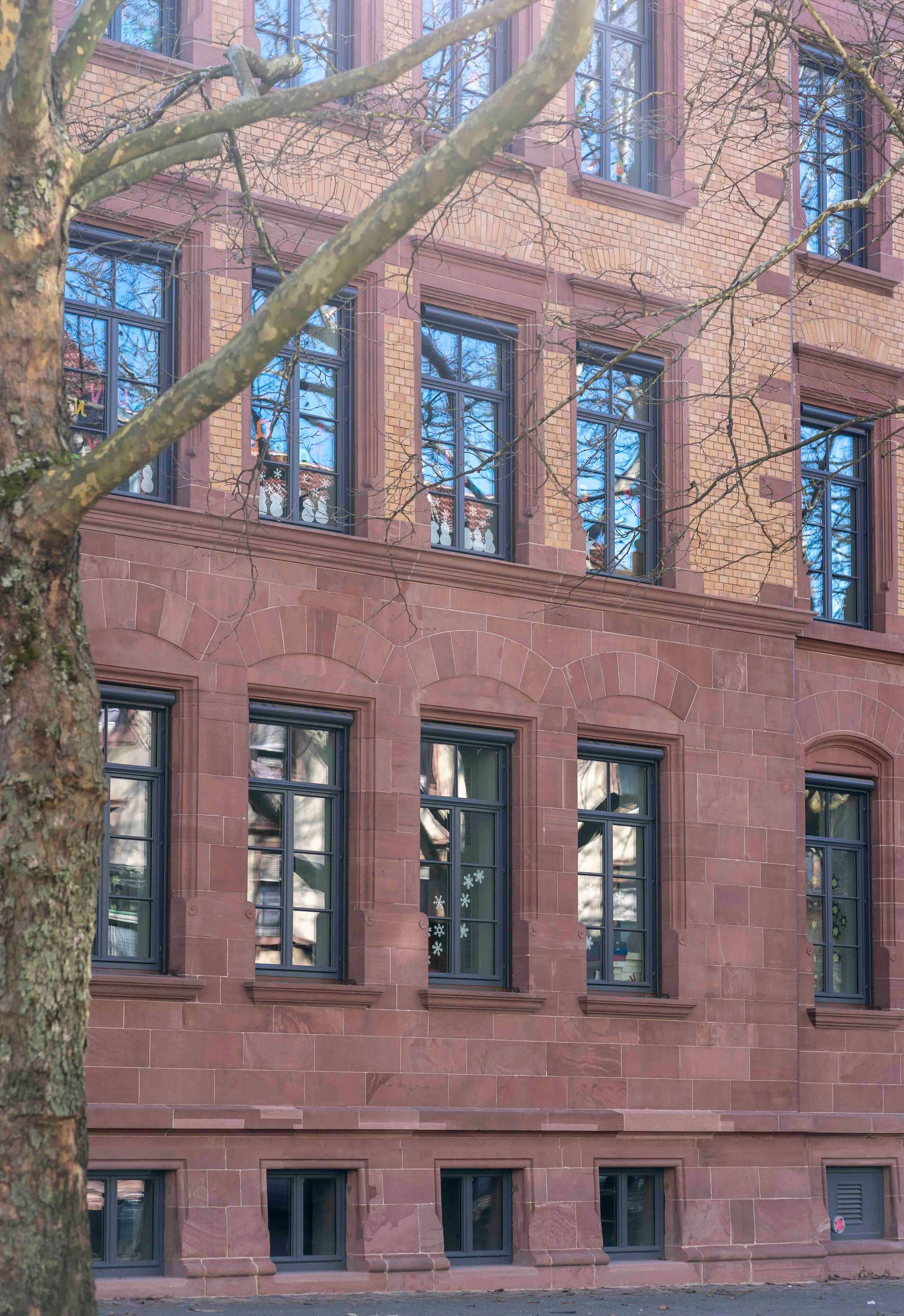 Rotes Backsteingebäude mit mehreren Fenstern; im Vordergrund ein Ast.