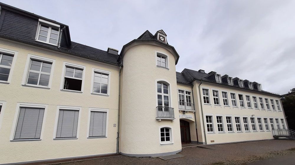 Cremefarbenes Schulgebäude mit Turm, vielen Fenstern und dunklem Dach unter bedecktem Himmel.