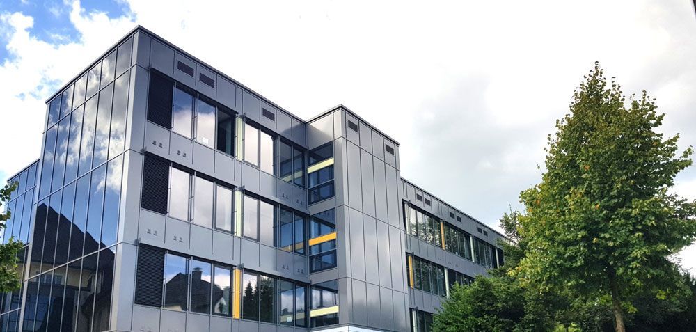 Modernes mehrstöckiges Gebäude mit Glasfassade und einem großen Baum im Vordergrund unter bewölktem Himmel.