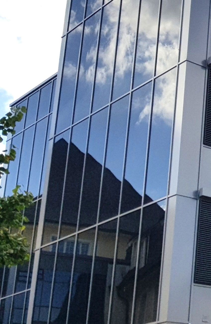 Modernes Gebäude mit reflektierender Glasfassade, Himmel und Nachbargebäude spiegeln sich darin.