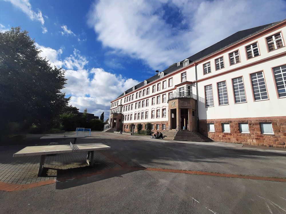 Großes Schulgebäude mit rotem Zierrand, bewölkter blauer Himmel und eine Tischtennisplatte im Vordergrund.