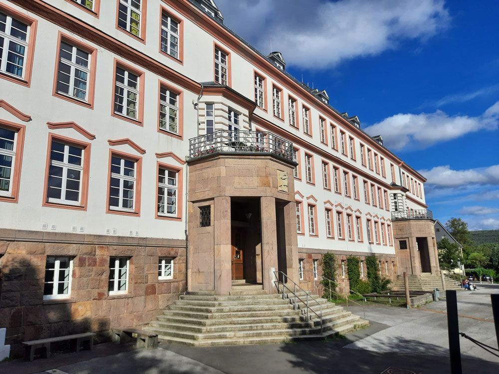 Mehrstöckiges, helles Gebäude mit vielen Fenstern, Steinfassade, Treppe zum Eingang, blauer Himmel.