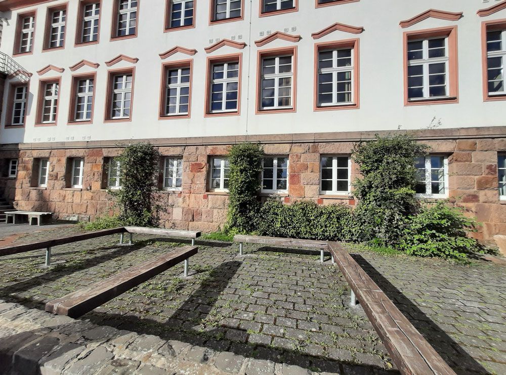 Außenansicht eines Steingebäudes mit Fenstern und Bänken in einem Innenhof. An der Fassade des Gebäudes wachsen einige Weinreben.