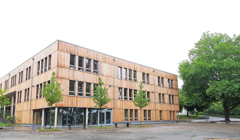 Modernes Holzgebäude mit vielen Fenstern, Bäumen und bewölktem Himmel.