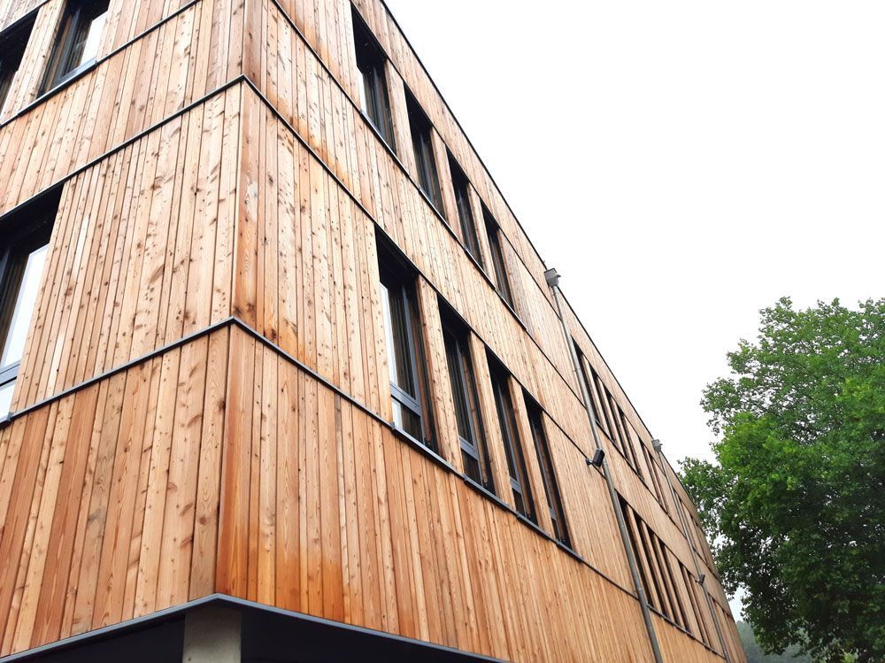 Ein mit Holz verkleidetes Gebäude mit Fensterreihen vor einem hellen Himmel, der teilweise von einem Baum verdeckt wird.