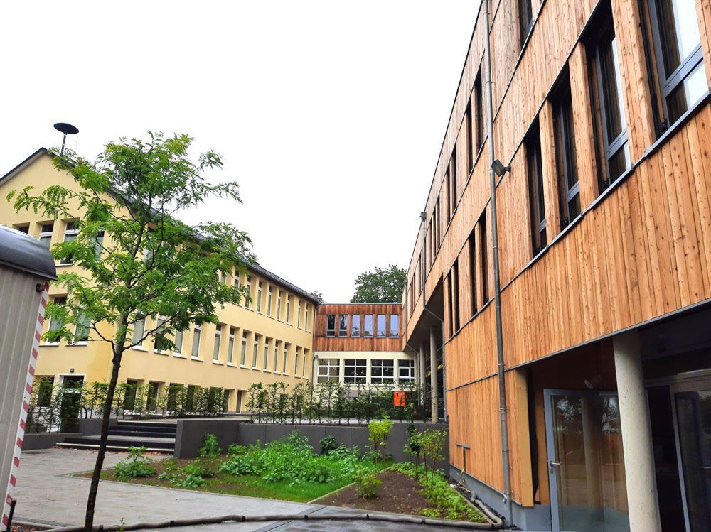 Schulhof mit modernem Holzgebäude und älterem gelbem Gebäude.
