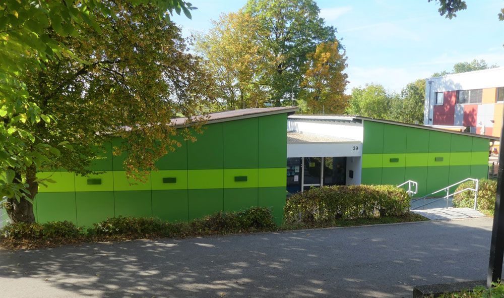 Grünes Gebäude mit hellgrünem Streifen, Eingang sichtbar. Bäume im Hintergrund. Sonniger Tag.