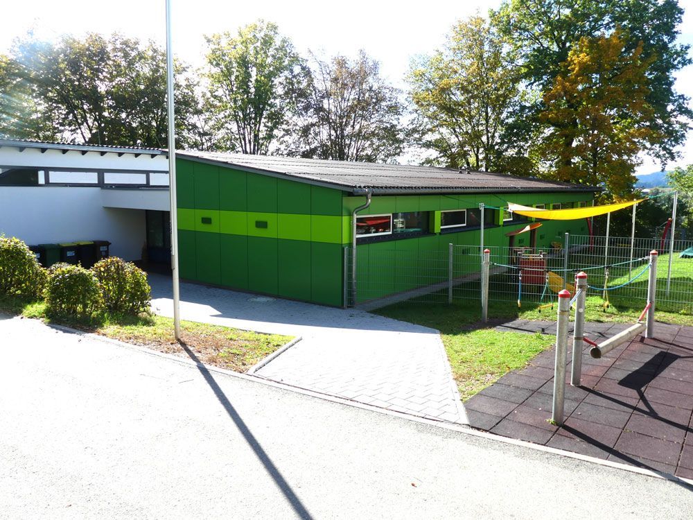 Grünes Gebäude mit schrägem Dach, neben einem Spielplatz mit schwarzem Gummibelag.