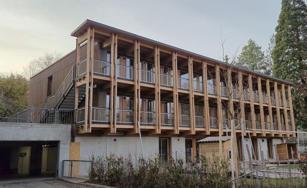 Dreigeschossiges Gebäude mit Holzrahmen, Balkonen und Betonfundament. Außentreppe und umliegende Bäume.