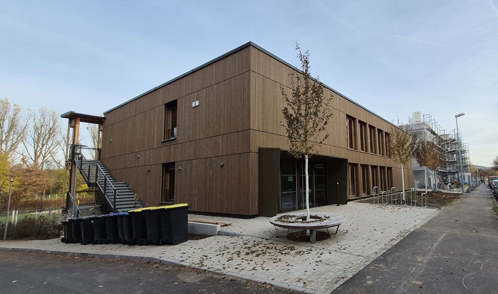 Braunes Holzgebäude mit Außentreppe und einem kleinen Baum davor.