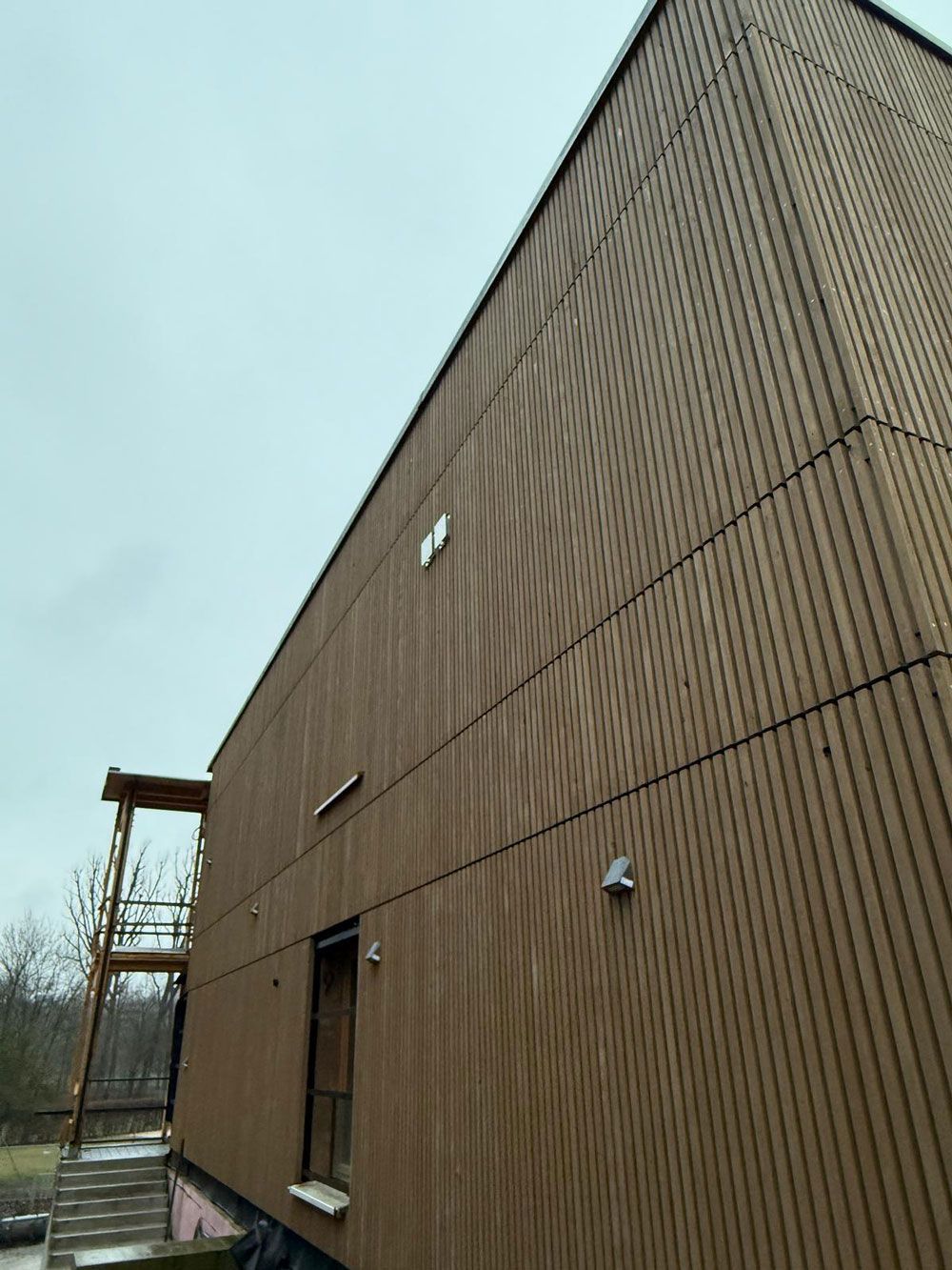 Braune Holzfassade mit vertikalen Linien. Mit kleiner Tür, Beleuchtung und Holztreppe. Bewölkter Himmel.
