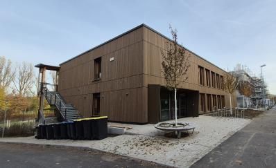 Braunes, zweistöckiges Gebäude mit Holzfassade, Eingang und Außentreppe. Davor befinden sich ein kleiner Baum und eine Kiesfläche.