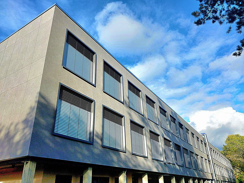 Graues, modernes Gebäude mit einer Fensterreihe, blauer Himmel mit Wolken im Hintergrund.