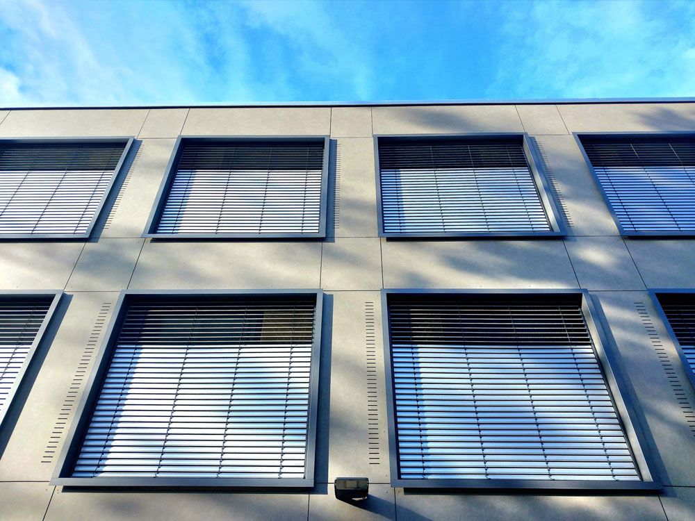 Moderne Gebäudefassade mit quadratischen Fenstern, horizontalen Jalousien und blauem Himmel im Hintergrund.