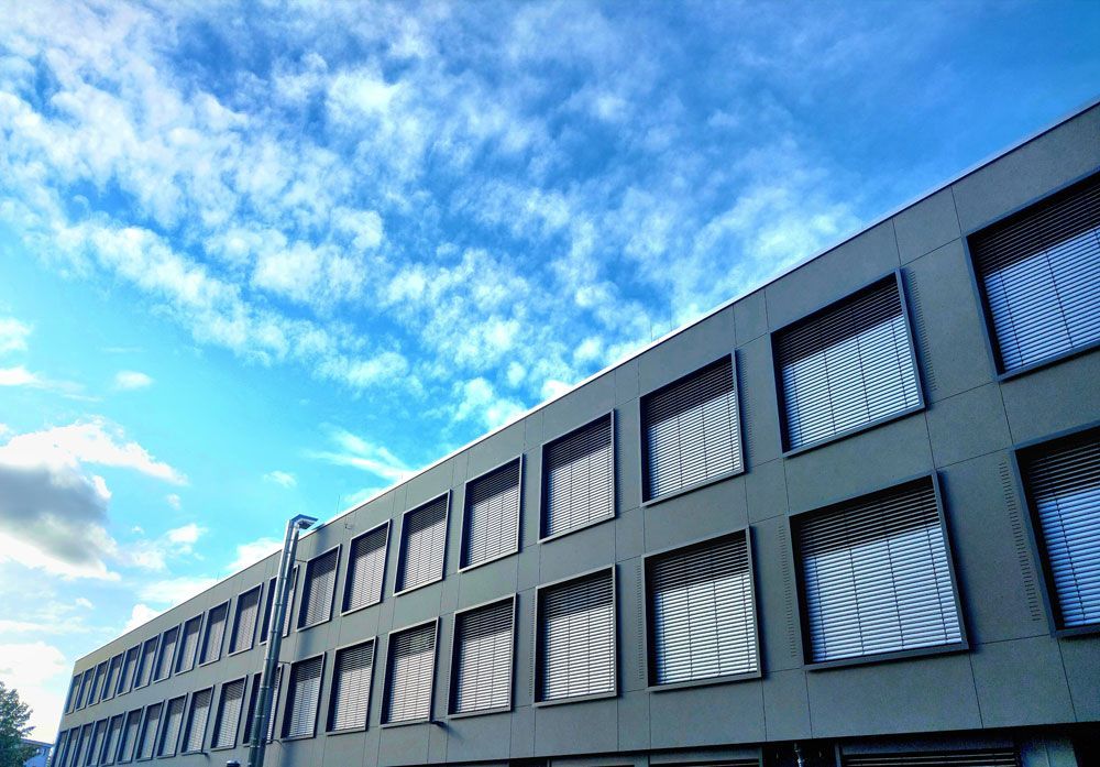 Graues Gebäude mit vielen quadratischen Fenstern vor einem strahlend blauen Himmel mit flauschigen weißen Wolken.