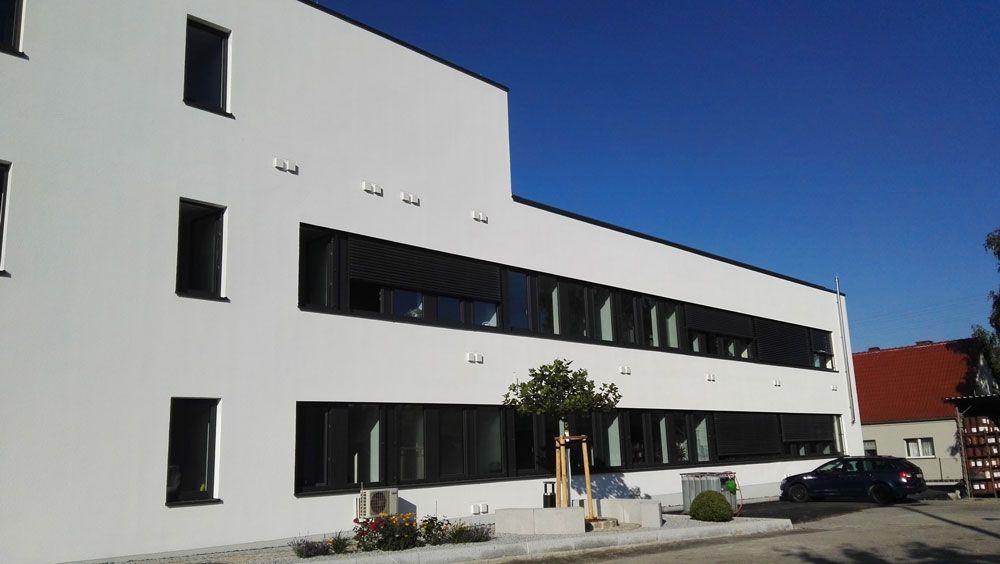 Weißes, modernes Gebäude mit dunklen Fenstern und Markisen, blauer Himmel, davor ein geparktes Auto.